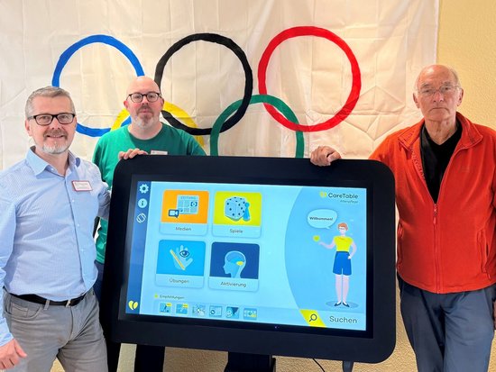 Drei Männer stehen vor einem Banner mit den olympischen Ringen und halten ein großes Tablet. Auf dem Bildschirm ist eine Benutzeroberfläche mit verschiedenen Symbolen, darunter ein Läufer, zu sehen.