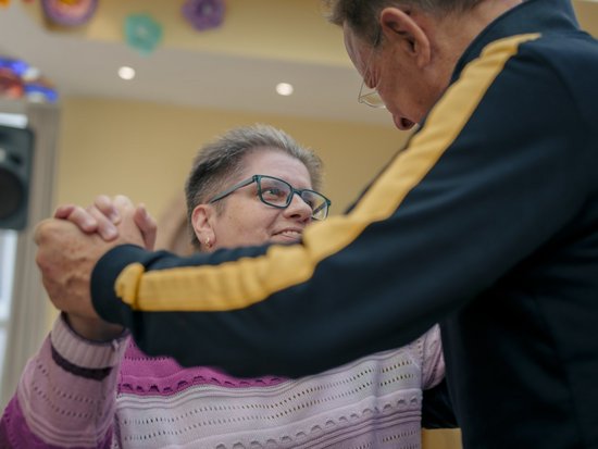 Ein älteres Paar tanzt gemeinsam. Die Frau mit Brille und rosa Pullover lächelt ihren Partner an, während er ihre Hände hält. Der Mann trägt eine dunkle Jacke mit gelbem Streifen.