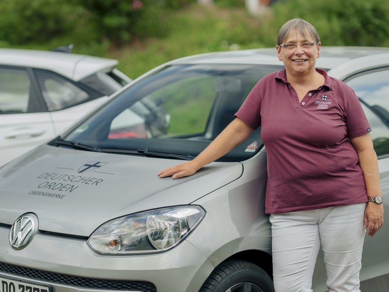 Eine Mitarbeiterin des ambulanten Pflegedienstes lehnt an ihrem Dienstfahrzeug und lächtelt in die Kamera.
