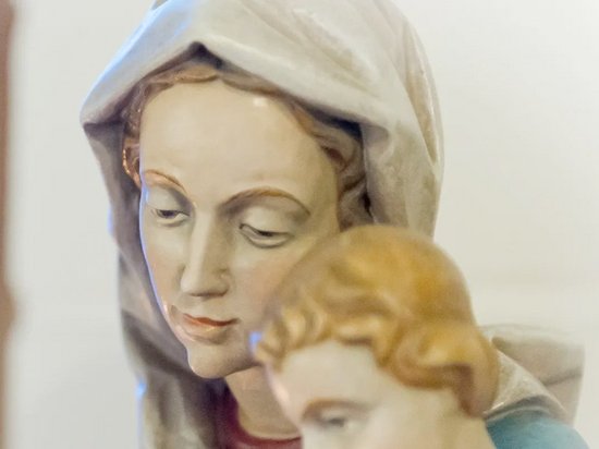 Der Kopf einer Marienstatue mit Jesuskind. Im Hintergrund ein blaues Kreuz in einem Fenster.